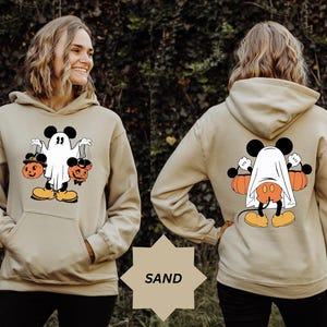 May include: Sand-colored hoodie featuring a Halloween-themed graphic. The front displays a cartoon character dressed as a ghost, holding two jack-o'-lanterns. The back shows the same character, partially covered by a sheet, with pumpkins on either side. The word "SAND" is visible.