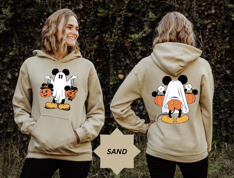 May include: Sand-colored hoodie featuring a Halloween-themed graphic. The front displays a cartoon character dressed as a ghost, holding two jack-o'-lanterns. The back shows the same character, partially covered by a sheet, with pumpkins on either side. The word "SAND" is visible.