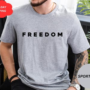 May include: Sport grey t-shirt with the word "FREEDOM" printed in black. The shirt is short-sleeved and made of a soft material. A red cloud-shaped sticker with the words "NEXT DAY SHIPPING" is in the upper left corner.