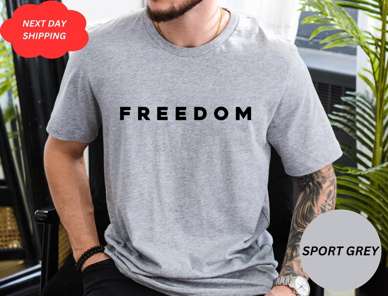 May include: Sport grey t-shirt with the word "FREEDOM" printed in black. The shirt is short-sleeved and made of a soft material. A red cloud-shaped sticker with the words "NEXT DAY SHIPPING" is in the upper left corner.