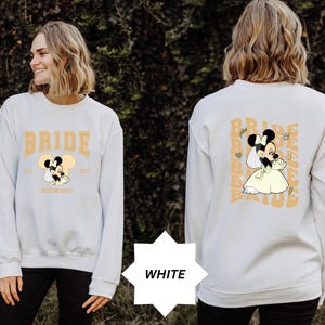 Può includere: Felpa bianca con un design di Minnie Mouse sposa. La parte anteriore mostra "BRIDE" in lettere dorate, l'anno "2024" e "Wedding Party". Il retro presenta un motivo ripetuto "BRIDE" con la grafica della sposa.