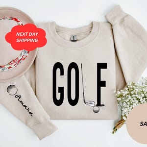 Pu&ograve; includere: Una felpa color sabbia con la parola "GOLF" stampata in nero. Un bastone da golf e una palla sono raffigurati accanto alla parola. La felpa ha una grafica di una palla da golf sulla manica.