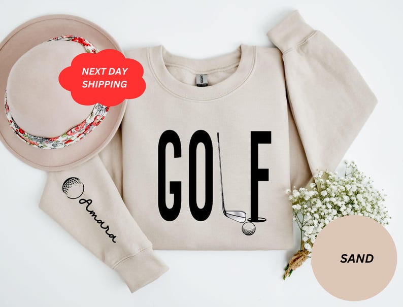 Pu&ograve; includere: Una felpa color sabbia con la parola "GOLF" stampata in nero. Un bastone da golf e una palla sono raffigurati accanto alla parola. La felpa ha una grafica di una palla da golf sulla manica.