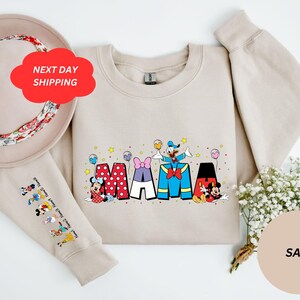 Puede incluir: Una sudadera beige con un gr&aacute;fico de Mickey Mouse, Minnie Mouse, Donald Duck y Goofy deletreando "MAMA" en letras de colores. La sudadera tiene mangas largas y cuello redondo. Las mangas tienen un estampado de varios personajes de Disney.