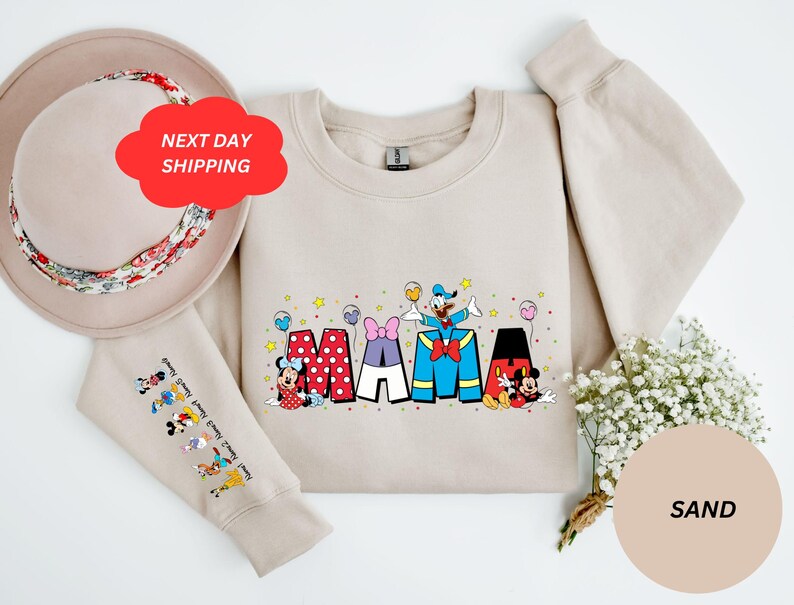 Puede incluir: Una sudadera beige con un gr&aacute;fico de Mickey Mouse, Minnie Mouse, Donald Duck y Goofy deletreando "MAMA" en letras de colores. La sudadera tiene mangas largas y cuello redondo. Las mangas tienen un estampado de varios personajes de Disney.