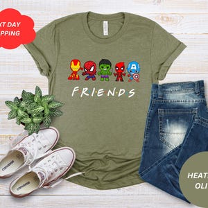 Puede incluir: Una camiseta verde con un gr&aacute;fico de cinco superh&eacute;roes de dibujos animados con la palabra "FRIENDS" debajo. La camiseta est&aacute; sobre una superficie de madera con un par de zapatillas blancas y una planta en maceta.