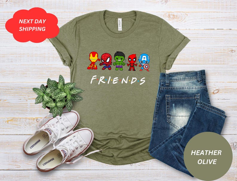 Puede incluir: Una camiseta verde con un gr&aacute;fico de cinco superh&eacute;roes de dibujos animados con la palabra "FRIENDS" debajo. La camiseta est&aacute; sobre una superficie de madera con un par de zapatillas blancas y una planta en maceta.