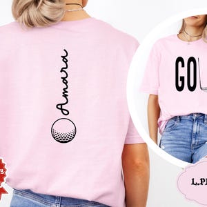 May include: Light pink t-shirts with golf-themed designs. One shirt features the word "GOLF" with a golf club and ball graphic. The other shirt has the name "Amara" vertically with a golf ball graphic.