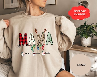 May include: A sand-colored sweatshirt with the word "MAMA" in a festive design. The "M" is red plaid, the first "A" has a green tree pattern, the second "A" has a leopard print. Below are snowmen with names. Christmas-themed apparel.