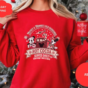 May include: Red Christmas sweatshirt with Mickey Mouse and Minnie Mouse holding a mug of hot cocoa. The text on the sweatshirt reads "Mickey & Minnie's Old Fashioned *Hot Cocoa* Served Daily Warm Up Here - Open All Winter".