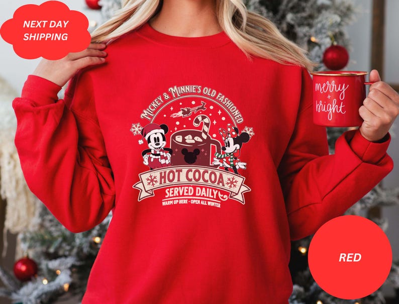 May include: Red Christmas sweatshirt with Mickey Mouse and Minnie Mouse holding a mug of hot cocoa. The text on the sweatshirt reads "Mickey & Minnie's Old Fashioned *Hot Cocoa* Served Daily Warm Up Here - Open All Winter".