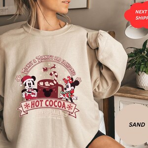 May include: A beige sweatshirt with a Christmas-themed design featuring Mickey and Minnie Mouse enjoying hot cocoa. The text reads "Mickey & Minnie's Old Fashioned Hot Cocoa".