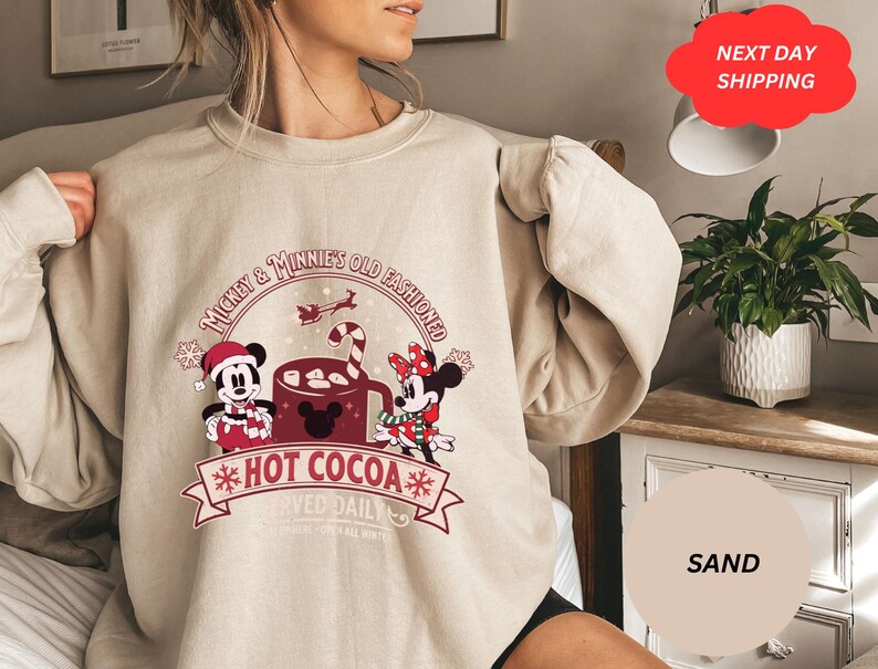 May include: A beige sweatshirt with a Christmas-themed design featuring Mickey and Minnie Mouse enjoying hot cocoa. The text reads "Mickey & Minnie's Old Fashioned Hot Cocoa".