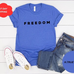 May include: A blue t-shirt with the word "FREEDOM" in black block letters. The shirt is paired with blue jeans and white sneakers with red accents. A red cloud-shaped graphic reads "NEXT DAY SHIPPING".