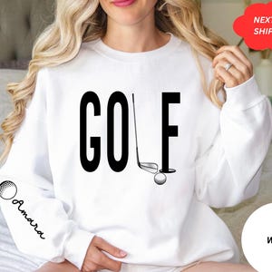 Pu&ograve; includere: Felpa bianca con testo nero che dice "GOLF" e una grafica di una mazza da golf e una palla. La felpa ha una grafica di una palla da golf sulla manica con la parola "Amara" scritta sotto.