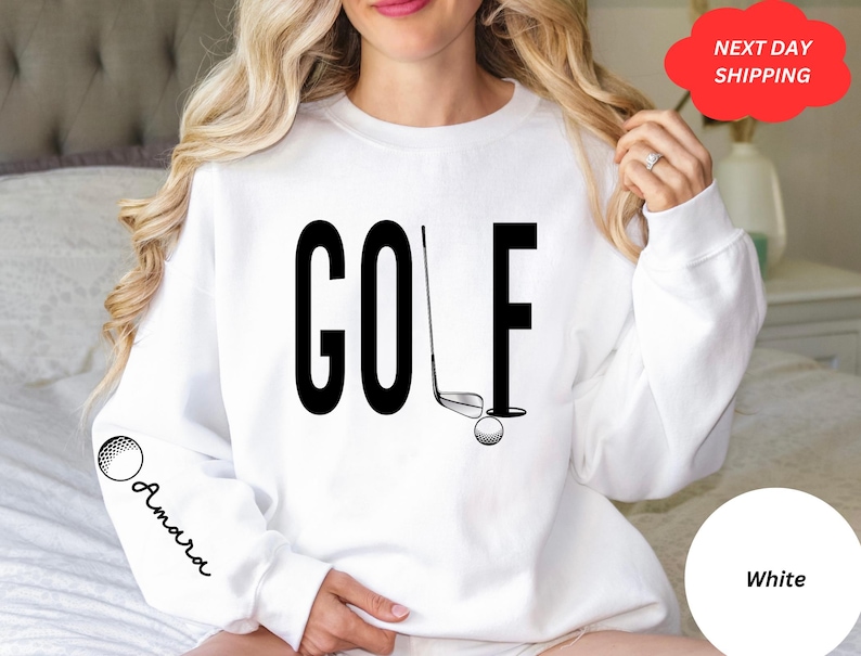 Pu&ograve; includere: Felpa bianca con testo nero che dice "GOLF" e una grafica di una mazza da golf e una palla. La felpa ha una grafica di una palla da golf sulla manica con la parola "Amara" scritta sotto.