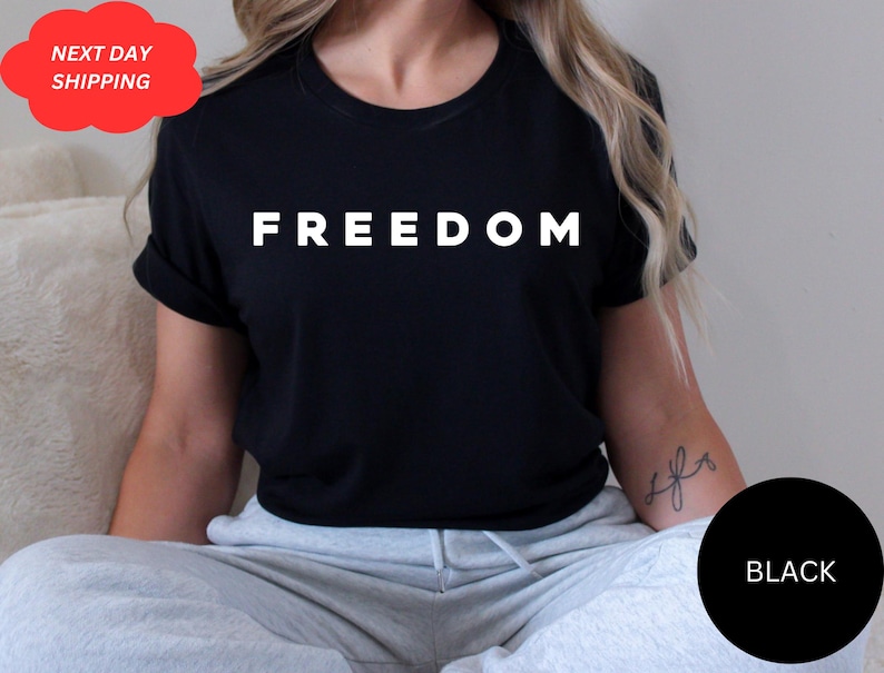 May include: Black t-shirt with the word "FREEDOM" printed in white. The shirt has short sleeves and a round neckline. A black circle with the word "BLACK" is in the lower right corner.