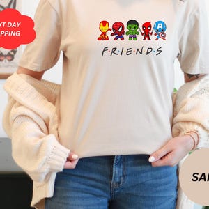 Puede incluir: Una camiseta blanca con un gr&aacute;fico de seis superh&eacute;roes de dibujos animados en fila. Los superh&eacute;roes son Iron Man, Spiderman, Hulk, Daredevil, Capit&aacute;n Am&eacute;rica y un personaje de la serie Friends. El texto "FRIENDS" est&aacute; impreso debajo de los superh&eacute;roes.