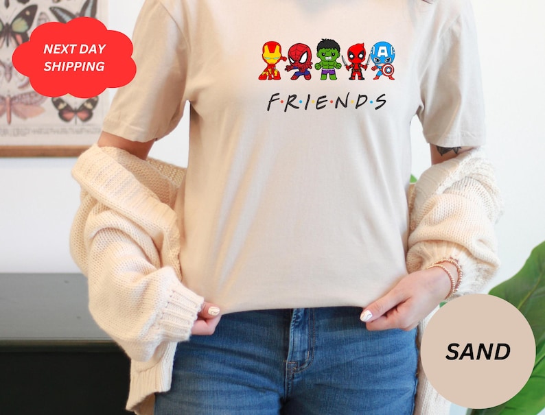 Puede incluir: Una camiseta blanca con un gr&aacute;fico de seis superh&eacute;roes de dibujos animados en fila. Los superh&eacute;roes son Iron Man, Spiderman, Hulk, Daredevil, Capit&aacute;n Am&eacute;rica y un personaje de la serie Friends. El texto "FRIENDS" est&aacute; impreso debajo de los superh&eacute;roes.