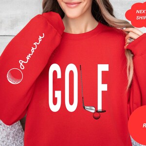 Pu&ograve; includere: Felpa rossa con la parola "GOLF" in lettere bianche e una grafica di una mazza da golf e una palla. La manica sinistra ha la parola "Amarra" in scrittura corsiva bianca.