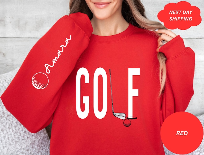 Pu&ograve; includere: Felpa rossa con la parola "GOLF" in lettere bianche e una grafica di una mazza da golf e una palla. La manica sinistra ha la parola "Amarra" in scrittura corsiva bianca.