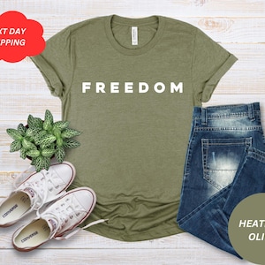 May include: An olive green t-shirt with the word "FREEDOM" in white block letters. The shirt is paired with blue jeans, white sneakers, and a small potted plant. The image includes a red cloud-shaped graphic with the text "NEXT DAY SHIPPING".