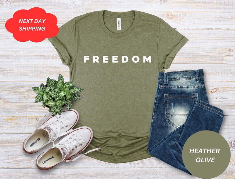 May include: An olive green t-shirt with the word "FREEDOM" in white block letters. The shirt is paired with blue jeans, white sneakers, and a small potted plant. The image includes a red cloud-shaped graphic with the text "NEXT DAY SHIPPING".