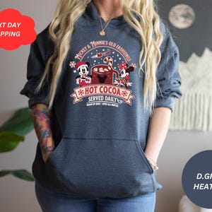 May include: A dark grey heather hoodie with a graphic of Mickey Mouse and Minnie Mouse holding a mug of hot cocoa. The graphic says "Mickey & Minnie's Old Fashioned *Hot Cocoa* Served Daily".