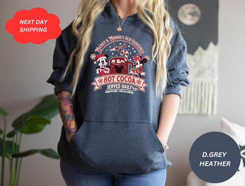 May include: A dark grey heather hoodie with a graphic of Mickey Mouse and Minnie Mouse holding a mug of hot cocoa. The graphic says "Mickey & Minnie's Old Fashioned *Hot Cocoa* Served Daily".