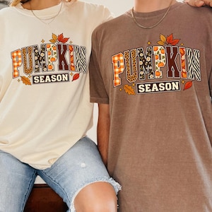 Può includere: Due magliette a maniche corte, una color crema e una marrone, con la scritta PUMPKIN SEASON in carattere varsity. Le lettere presentano motivi come quadretti arancioni, stampa leopardata, fiori e strisce zebrate, accentuati da piccole foglie autunnali.