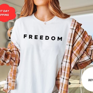 May include: White t-shirt with the word "FREEDOM" printed in black. The person is wearing a brown and pink plaid shirt over the white t-shirt. A white circle with the word "WHITE" is in the bottom right corner.