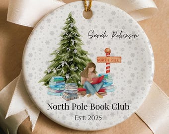 May include: A round ceramic Christmas ornament with a gold string, featuring a watercolor illustration of a Christmas tree, a person reading, and a sign that says "North Pole". The ornament is on a wrapped gift with a white ribbon.