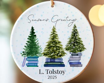 May include: A round Christmas ornament with a gold string. The ornament features three watercolor Christmas trees atop stacks of books. The text "Seasons Greetings", "L. Tolstoy", and "2025" are printed on the ornament.