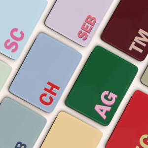 May include: A collection of rectangular, personalized items in various colors, including blue, green, and red. Each item features a two-letter or three-letter initial in a contrasting color, such as pink or white, creating a custom design.
