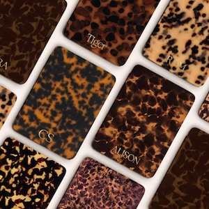 May include: A collection of rectangular phone cases with a tortoiseshell pattern in shades of brown, tan, and black. Some cases have gold lettering with names like "SARA" and "ALISON". The cases are arranged on a white surface.