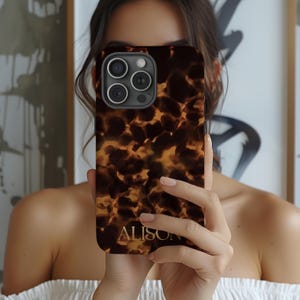 May include: A brown and gold tortoiseshell-patterned phone case with the name "ALISON" in gold lettering. The case has a camera cutout with three lenses. The phone case is held in front of a person.
