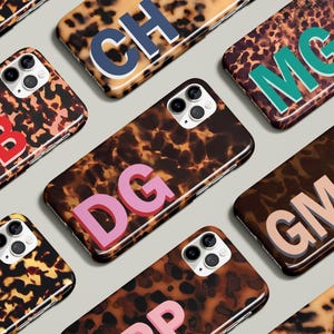 May include: A collection of smartphone cases with a tortoiseshell pattern. Each case features a different set of initials in bold, contrasting colors, including red, pink, teal, and blue. The cases are designed for a modern aesthetic.