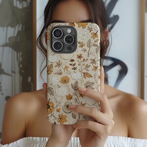Peut inclure: Coque de téléphone beige avec un motif floral sur le thème de l'automne, comprenant des citrouilles, des feuilles et des fleurs dans des tons de blanc, de marron et d'or. La coque a une finition brillante et une découpe pour l'appareil photo.