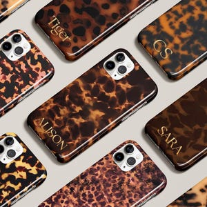 May include: A collection of tortoiseshell-patterned phone cases. The cases feature a variety of brown and tan hues, with some displaying personalized gold lettering. The cases are designed to fit a smartphone with a multi-lens camera.