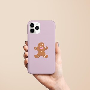 May include: A lilac phone case featuring a gingerbread man design. The gingerbread man is brown with a pink bow tie. The phone case is held in a hand against a neutral background.
