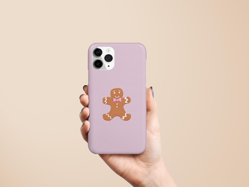 May include: A lilac phone case featuring a gingerbread man design. The gingerbread man is brown with a pink bow tie. The phone case is held in a hand against a neutral background.