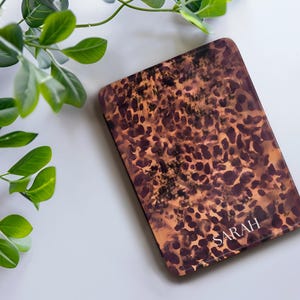 K&ouml;nnte beinhalten: Eine rechteckige H&uuml;lle mit Leopardenmuster in Braun- und Beiget&ouml;nen. Das Wort "SARAH" ist in Wei&szlig; in der unteren rechten Ecke aufgedruckt. Gr&uuml;ne, belaubte Zweige befinden sich in der oberen linken Ecke.