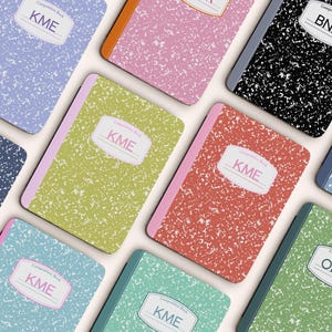 May include: A collection of composition notebooks in various colors, including blue, pink, green, and black. Each notebook features a speckled cover and a label with initials or a name. The notebooks are arranged in a visually appealing pattern.