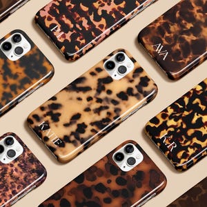 May include: A collection of tortoiseshell-patterned phone cases. The cases feature a glossy finish and are in shades of brown, tan, and black. Some cases have personalized initials in white. The cases are designed for a smartphone.