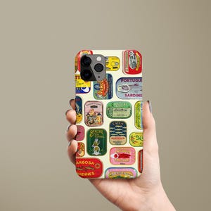Może przedstawiać: Etui na telefon z kolażem etykiet po puszkach sardynek w stylu vintage w r&oacute;żnych kolorach. Etykiety zawierają tekst i ilustracje na kremowym tle. Etui na telefon jest trzymane w dłoni.
