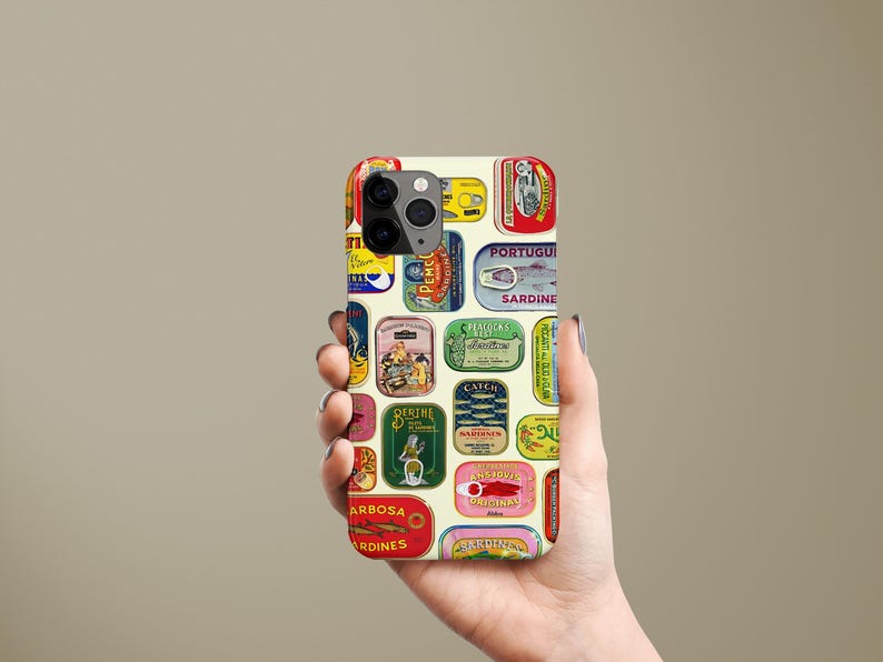 Może przedstawiać: Etui na telefon z kolażem etykiet po puszkach sardynek w stylu vintage w r&oacute;żnych kolorach. Etykiety zawierają tekst i ilustracje na kremowym tle. Etui na telefon jest trzymane w dłoni.