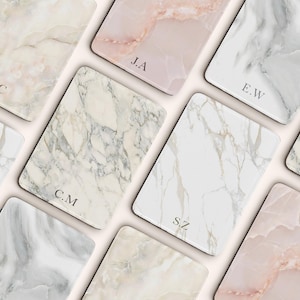 May include: A collection of rectangular marble-patterned trays in various colors, including white, pink, and gray. Each tray features a unique veining pattern and is personalized with initials in a subtle font. The trays are arranged on a light background.