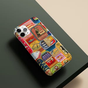 Może przedstawiać: Etui na smartfon z kolażem etykiet z puszek po sardynkach w stylu vintage. Etykiety są w r&oacute;żnych kolorach, w tym czerwonym, ż&oacute;łtym, niebieskim i zielonym, z tekstem i ilustracjami. Etui leży na ciemnozielonej powierzchni.