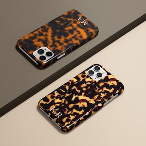 May include: Two tortoise shell patterned phone cases with white initials. The cases are black and brown with a glossy finish. The cases are on a neutral background with a diagonal dark brown line.