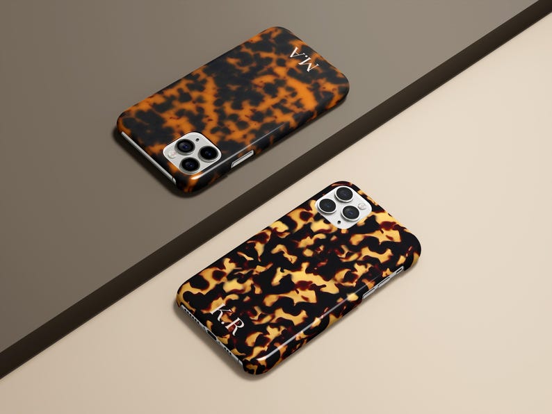 May include: Two tortoise shell patterned phone cases with white initials. The cases are black and brown with a glossy finish. The cases are on a neutral background with a diagonal dark brown line.